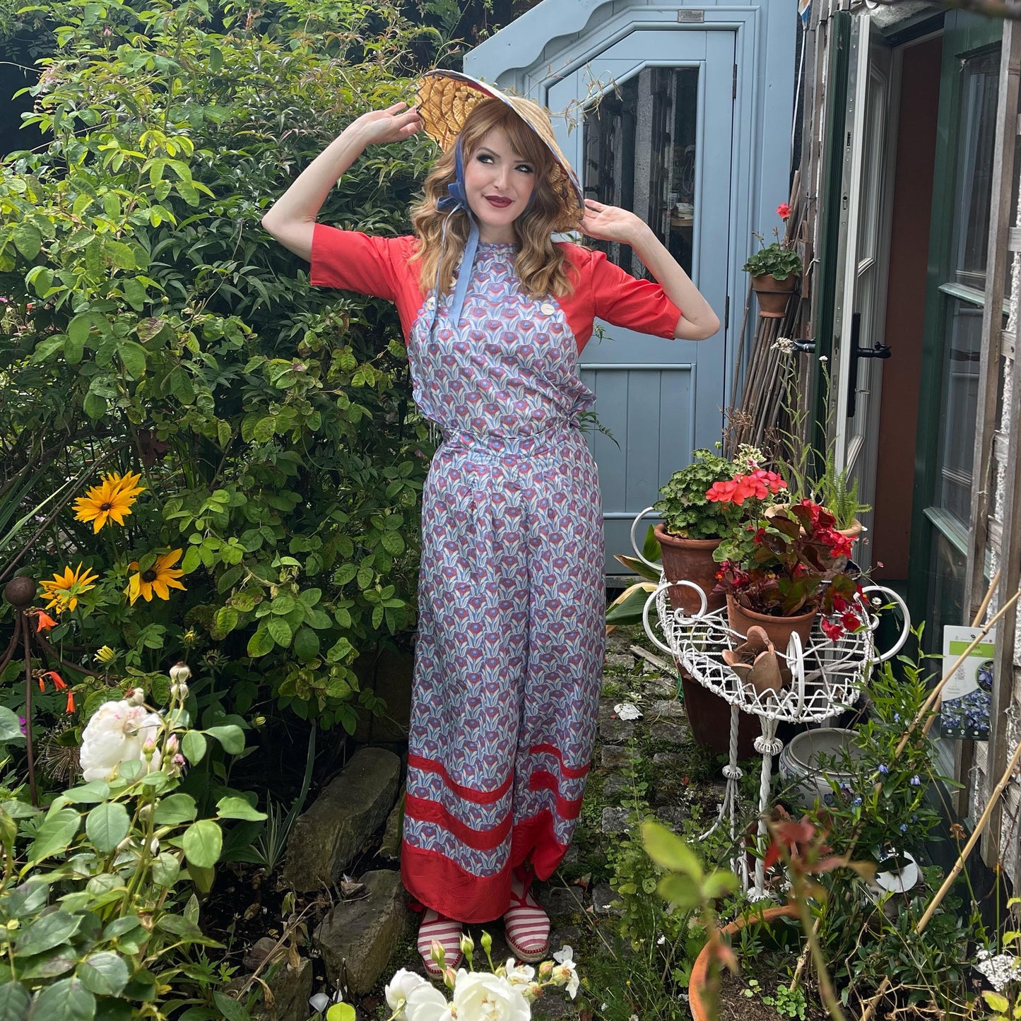 Samantha wearing colour block 1930s beach pajamas with straw hat standing in cottage garden