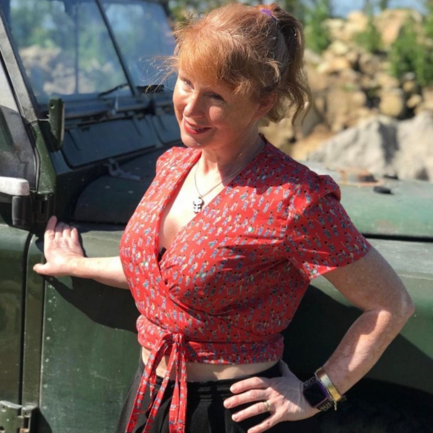 Woman in a red patterned top standing next to a green vehicle in a natural setting