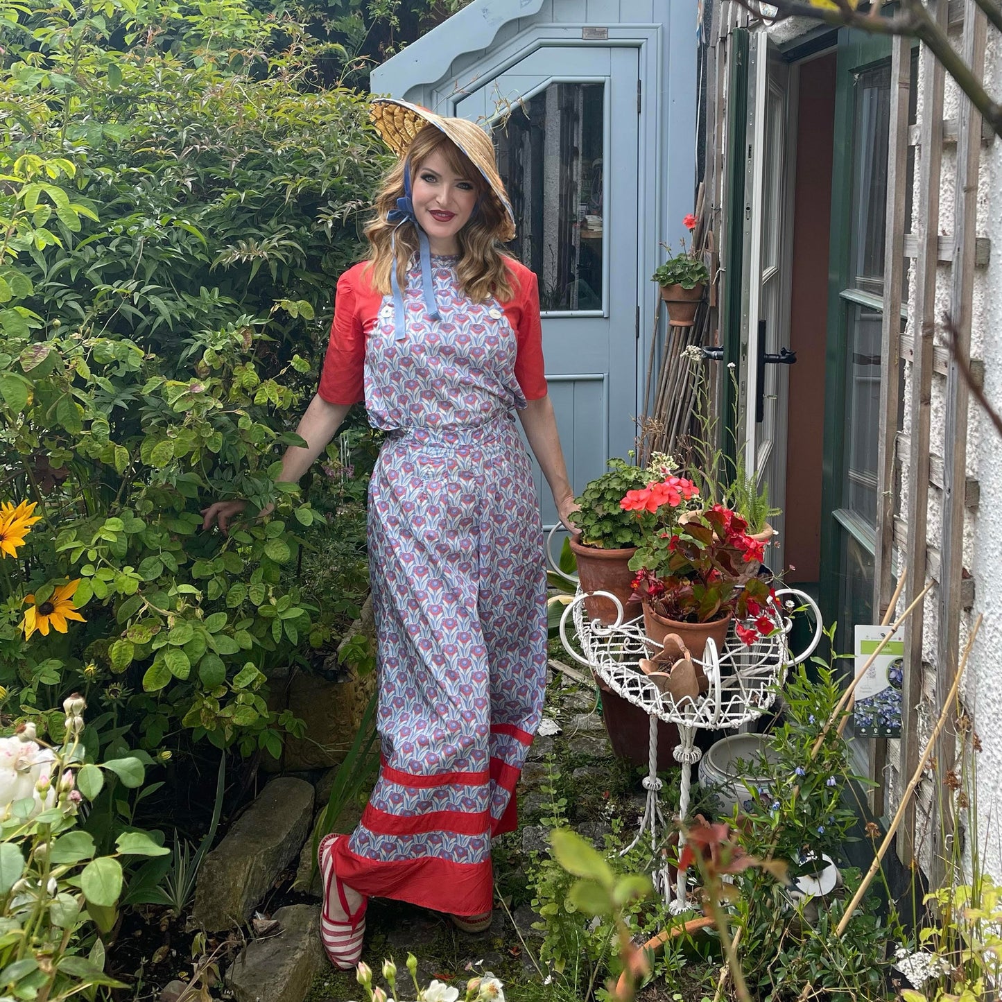 Samantha wearing colour block 1930s beach pajamas with straw hat standing in cottage garden 