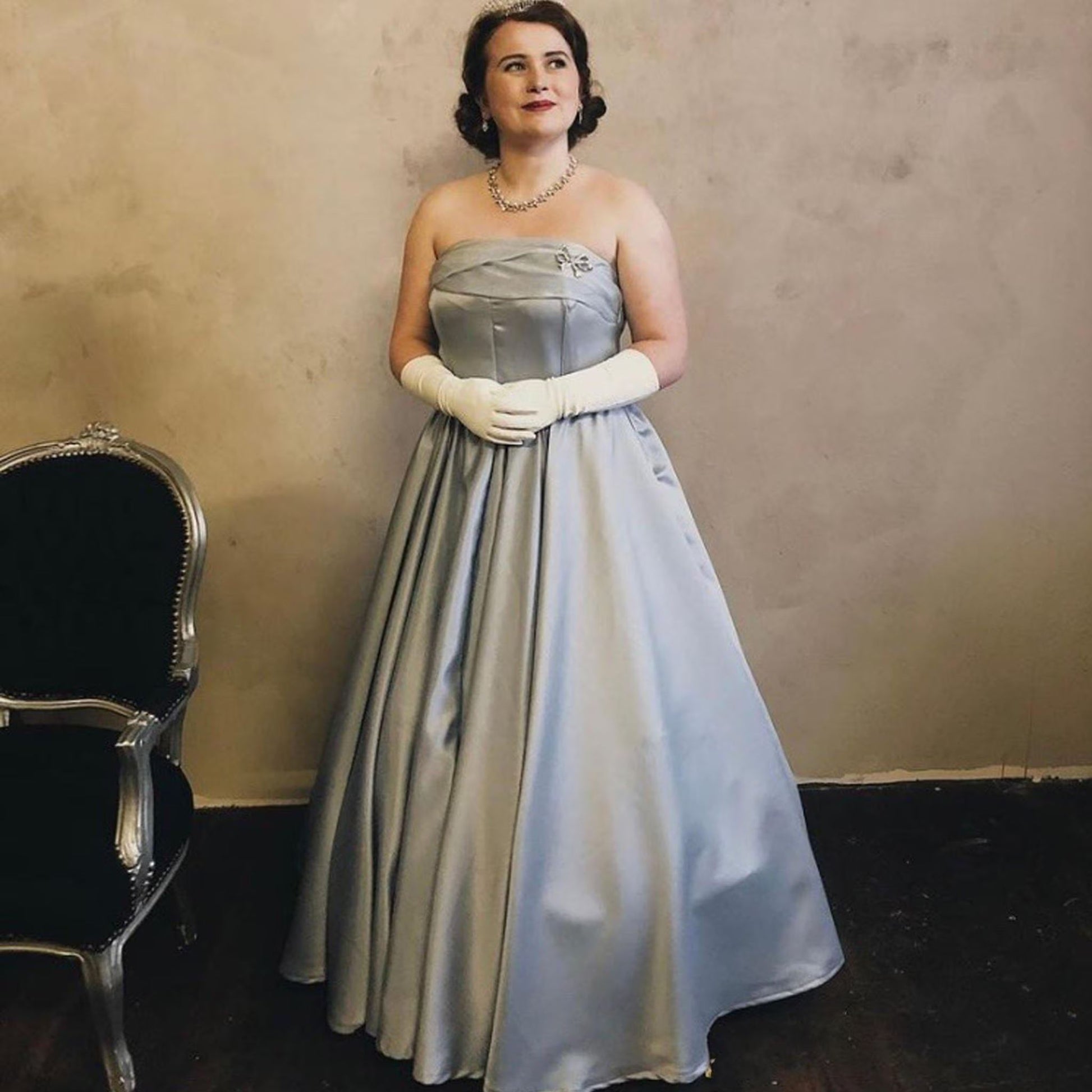 A strapless, floor-length light blue gown with a fitted bodice and a full, pleated skirt. The ensemble includes white elbow-length gloves, a silver-toned necklace, and a small silver brooch pinned to the upper left side of the bodice.