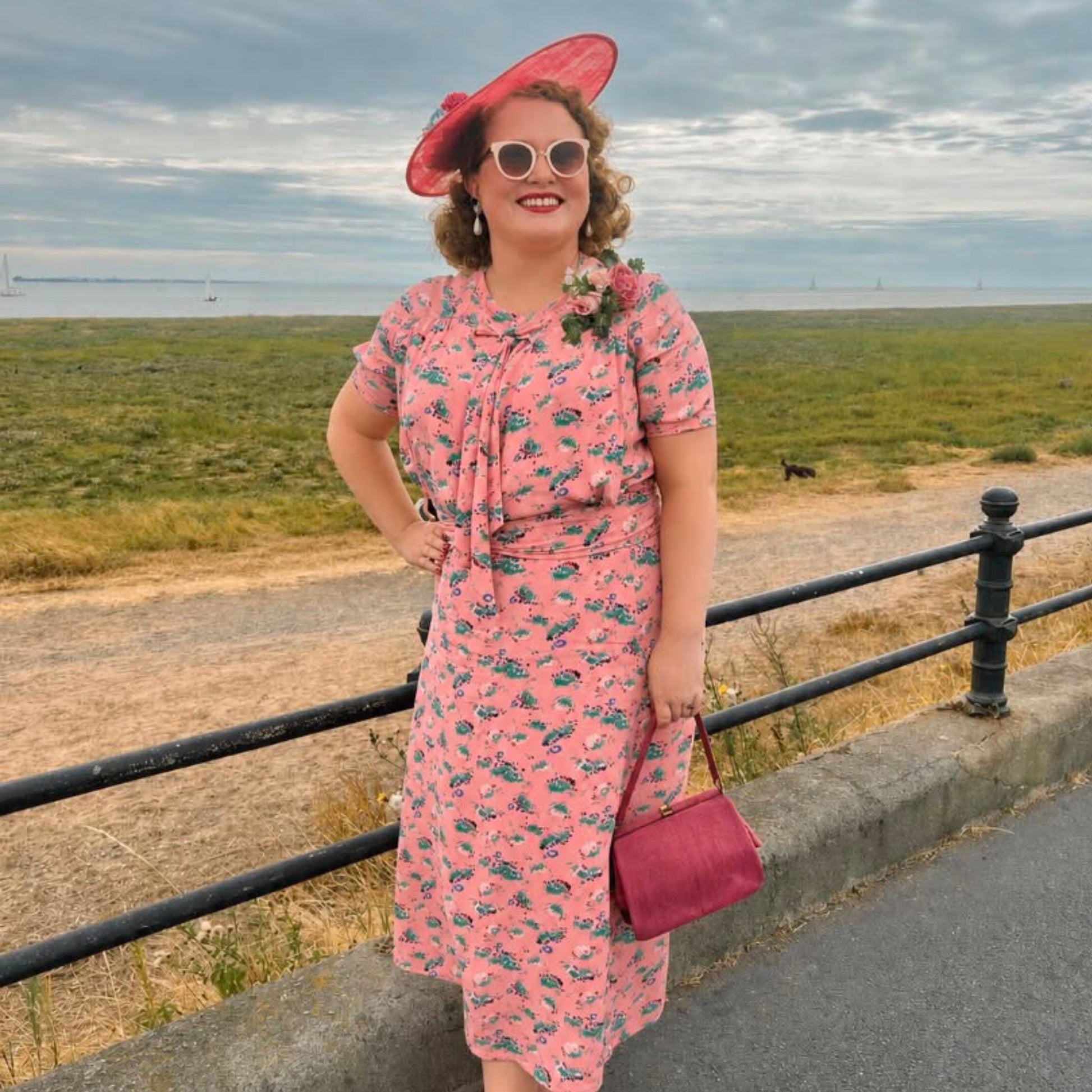 A woman stands outdoors by a railing wearing a pink floral midi dress with a matching wide-brim hat, sunglasses, and a small pink handbag, with a grassy field and cloudy sky behind her.