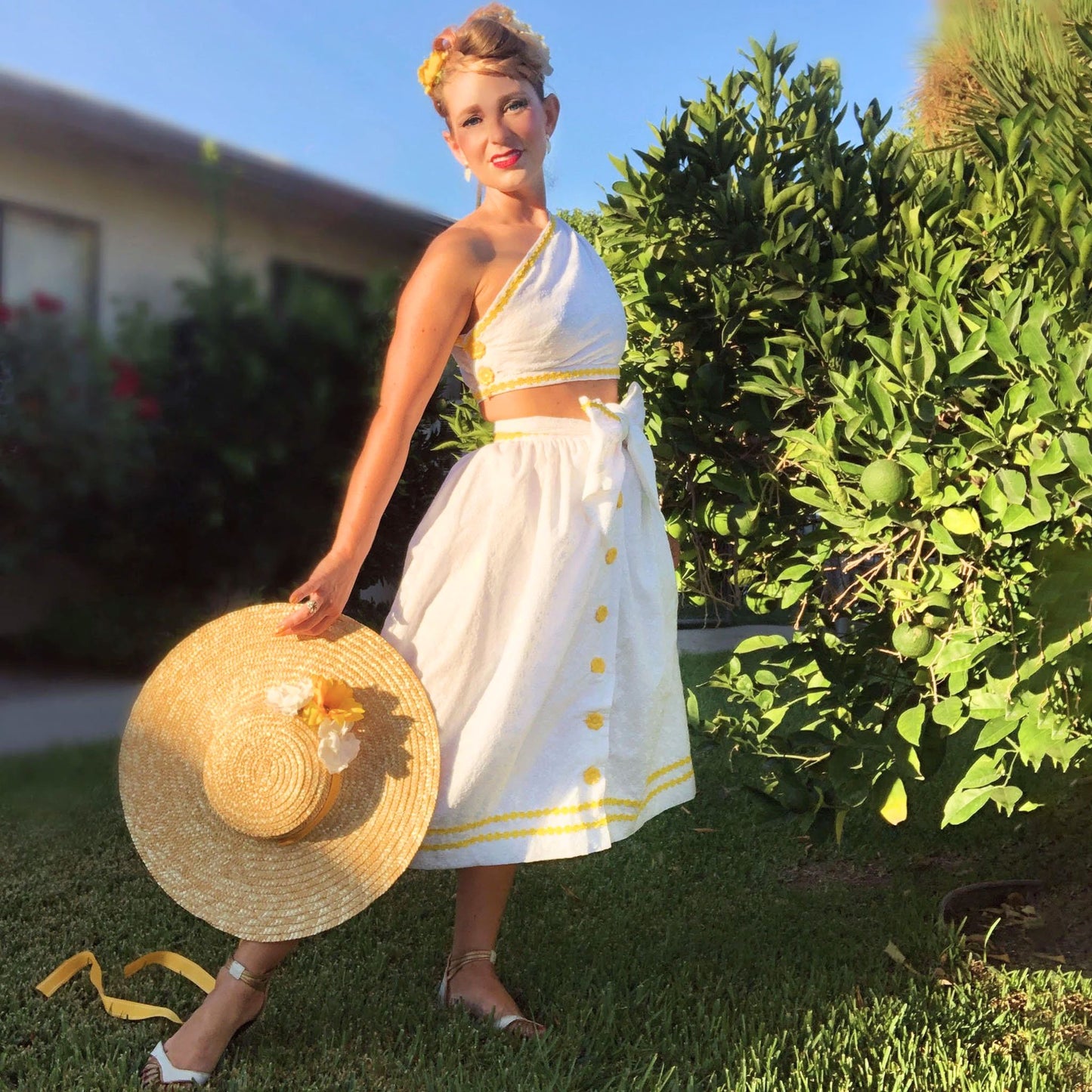 Woman in a white dress with yellow buttons holding a straw hat outdoors.