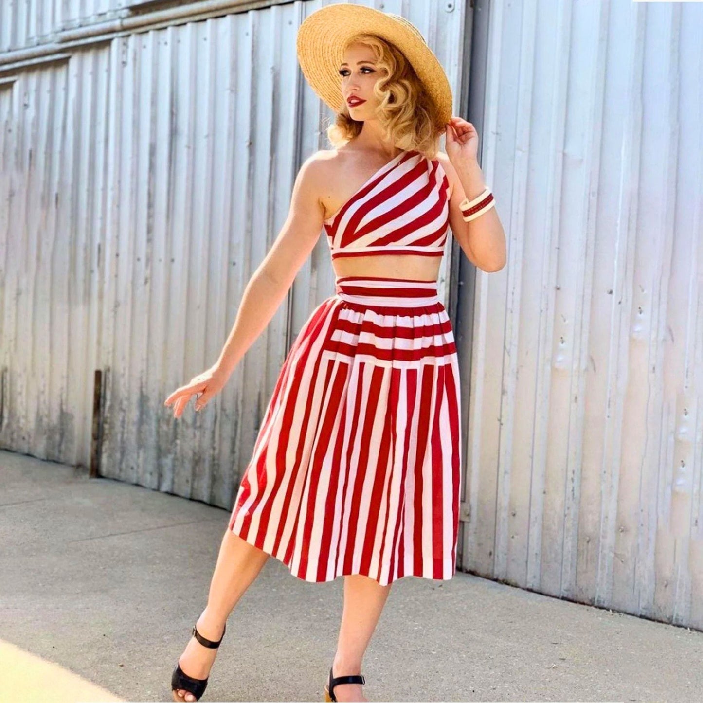 Woman wearing a red and white striped dress with a straw hat against a corrugated metal wall.