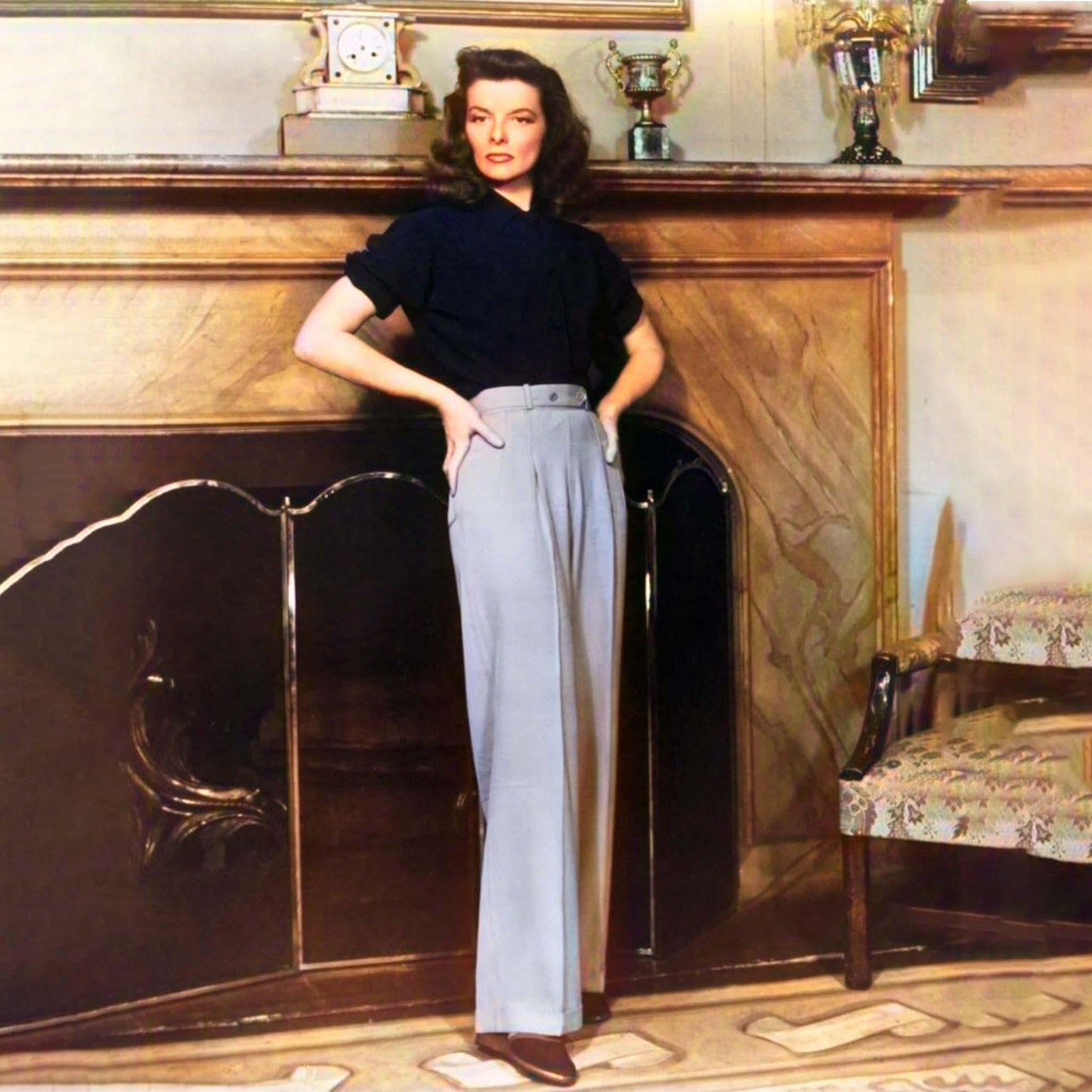 Woman in a black top and light gray pants standing in a room with a fireplace.