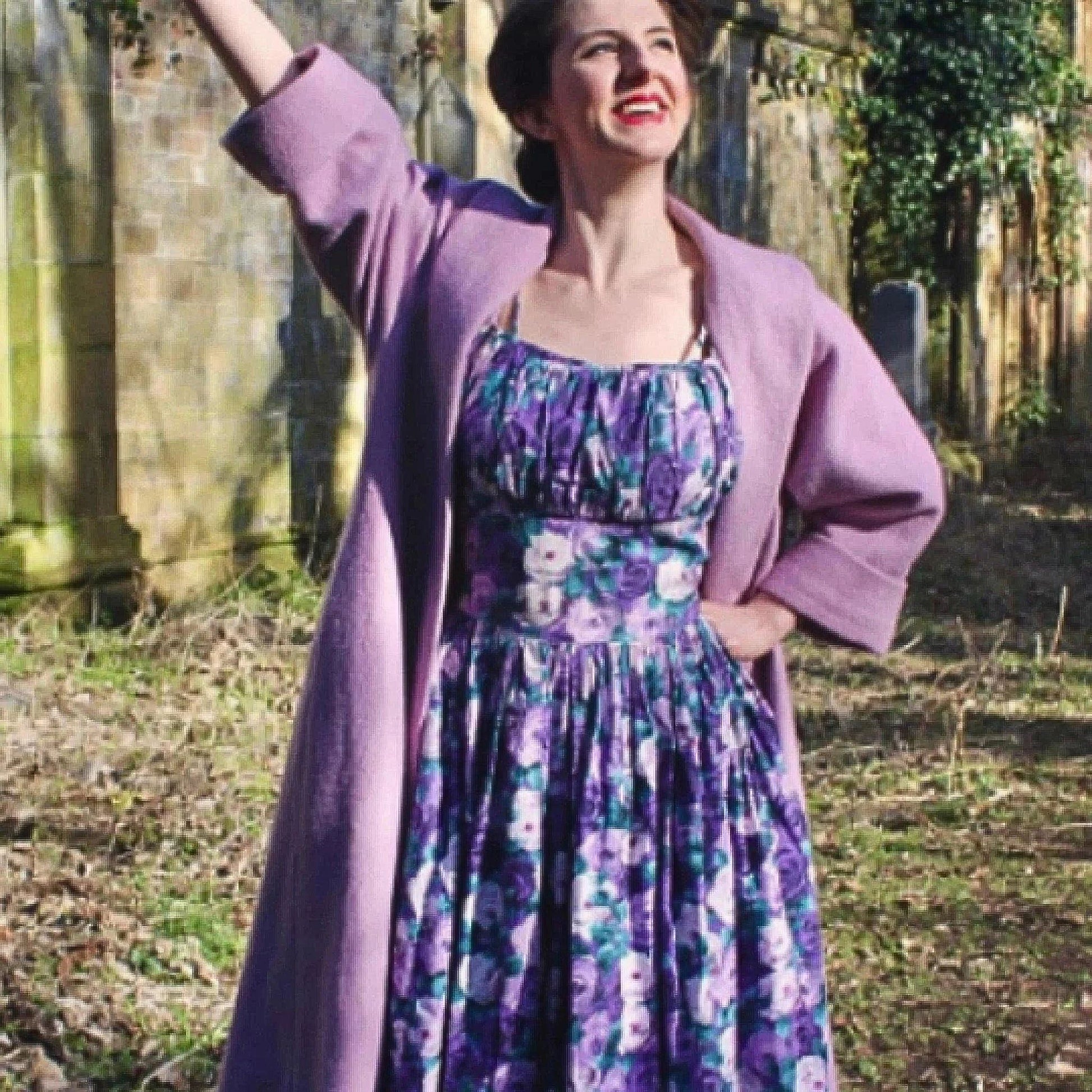 Woman in a floral dress and purple cardigan standing outdoors with a stone wall and greenery in the background.