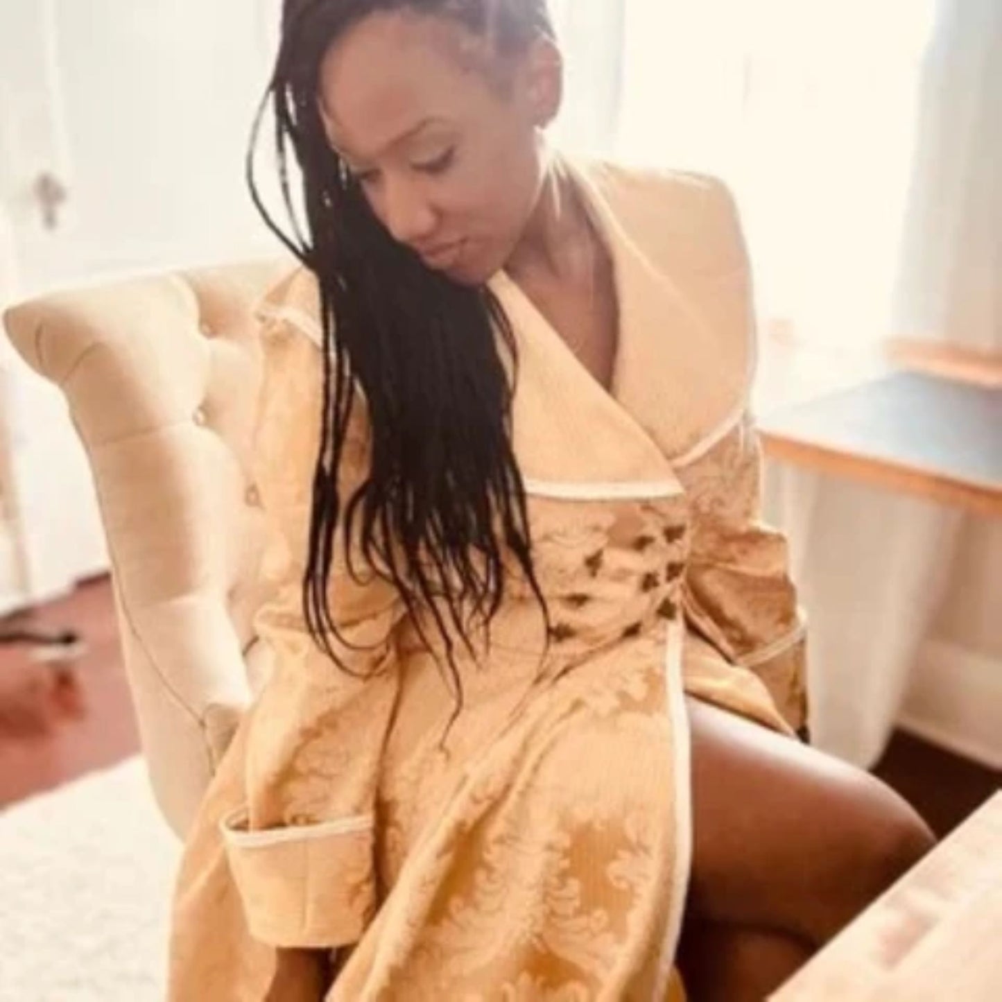 A woman with long dark braids sits in a cream tufted armchair, wearing a beige patterned robe in a brightly lit indoor setting.