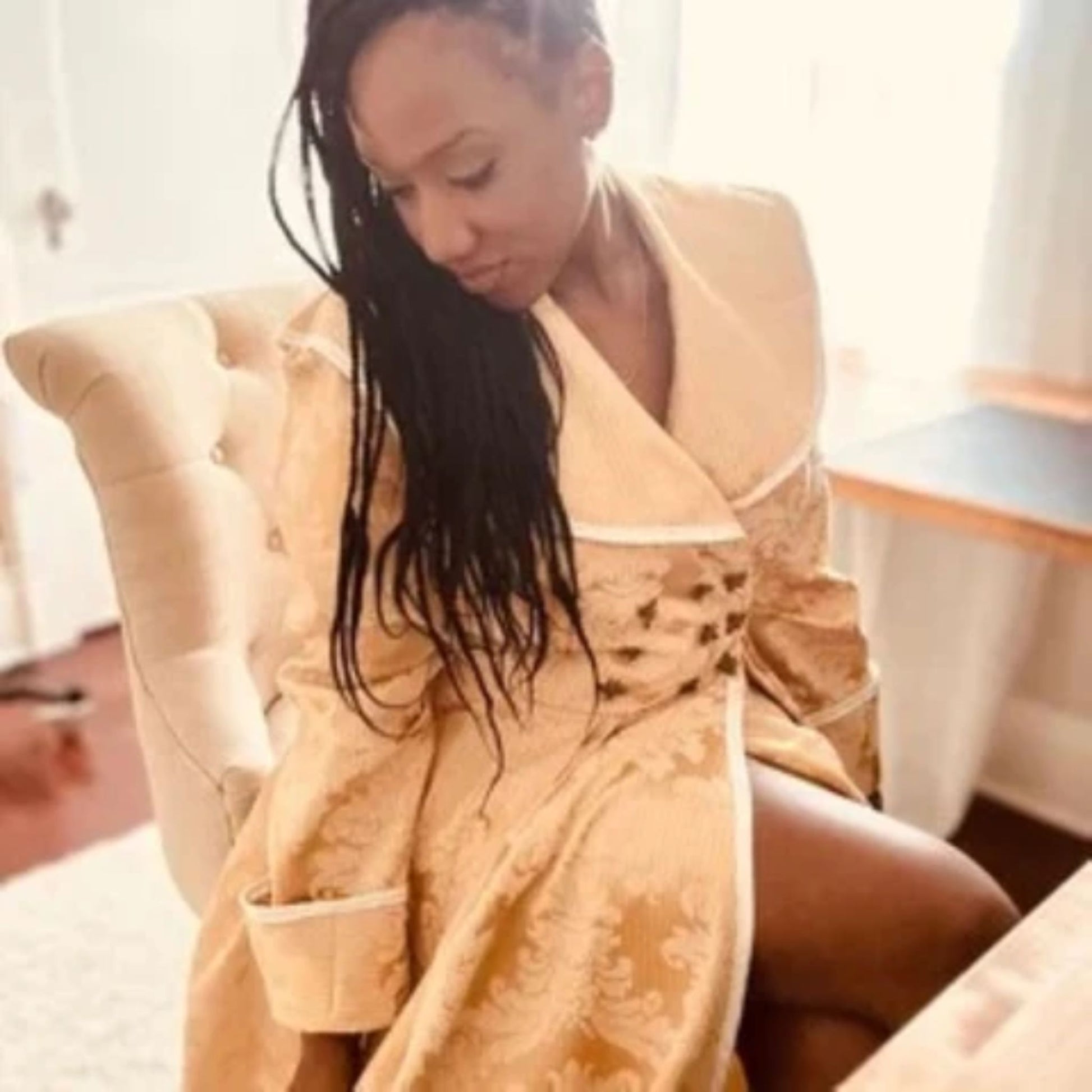 A woman with long dark braids sits in a cream tufted armchair, wearing a beige patterned robe in a brightly lit indoor setting.