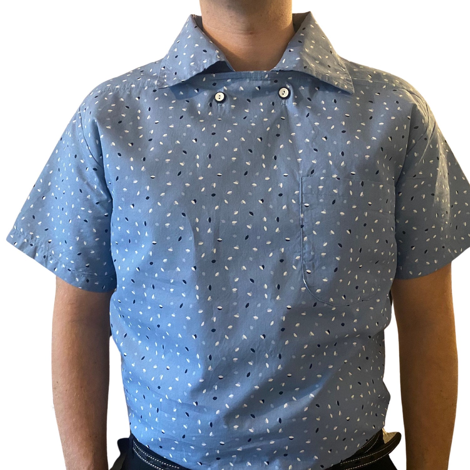 Man wearing blue 1950s sports shirtt with white and blue fleck print. Shirt has collar and two button neck fastening with short sleeves and chest pocket.